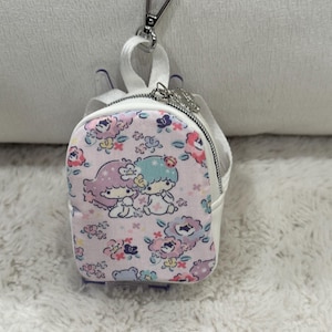 May include: A small, white and pink backpack-shaped keychain with a silver clasp. The front of the backpack features a floral pattern with cartoon characters. The zipper is silver, and the straps are white.