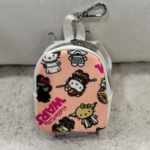 May include: A small, pink and white backpack-shaped keychain featuring Hello Kitty characters dressed as Star Wars characters. The keychain has a silver clasp and a white zipper. The words "Hello Kitty Wars" are printed on the front.