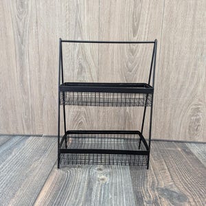 May include: Black metal two-tiered shelf with wire baskets. The shelf is freestanding and has a simple, modern design.