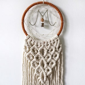 May include: A white macrame dream catcher with a brown wooden hoop and a small glass vial hanging from it. The vial is filled with brown and white materials.