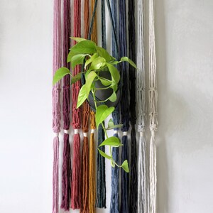 May include: A set of 12 macrame plant hangers in a variety of colours, including pink, red, orange, yellow, green, blue, and white. The hangers are made from cotton cord and have a simple design. A small potted plant hangs from one of the hangers.