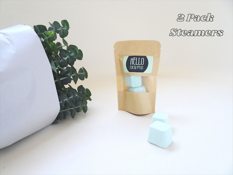 May include: A kraft paper bag with a clear window contains light blue cube-shaped bath steamers. The label reads "HELLO EUCALYPTUS". Two additional steamers are in front of the bag. The text "2 Pack Steamers" is above the bag. Green eucalyptus leaves are in the background.