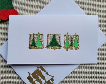 May include: White greeting card with three rectangular designs featuring green Christmas trees with gold glitter accents. The card is inside a white envelope, with a felt holly and berry decoration in the background.