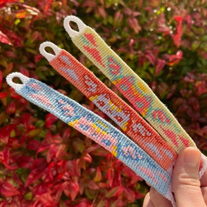 Puede incluir: Tres pulseras de la amistad con patrones tejidos coloridos y las palabras "Be Kind", "Happy" y "Good Vibes" en un esquema de colores rosa claro, naranja y azul.