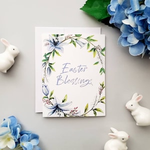 May include: A white card with a floral wreath border in shades of blue, green, and pink. The text "Easter Blessings" is written in purple inside the wreath.