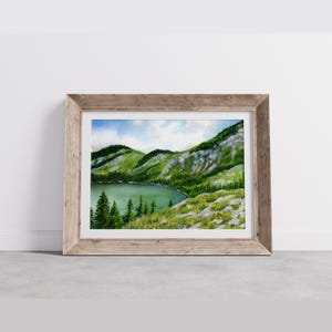 May include: Watercolor painting of a mountain lake scene, framed in a light brown wooden frame. The artwork depicts a serene lake surrounded by green mountains and trees under a blue sky with clouds. The painting evokes a sense of tranquility.