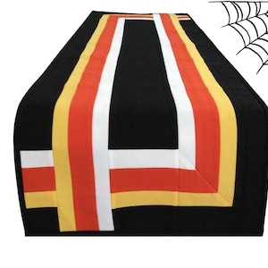 Può includere: Runner da tavolo nero di Halloween con un design a caramelle di mais. Il runner presenta una striscia centrale bianca, arancione e gialla, su sfondo nero. Un disegno di ragnatela è nell'angolo in alto a destra.