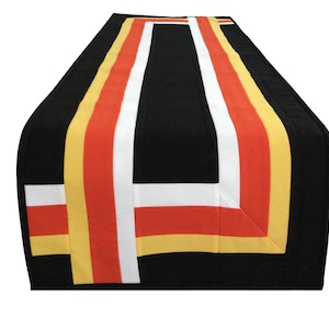 May include: A rectangular table runner featuring a solid black base with a geometric pattern of vertical and horizontal stripes in white, orange, and yellow. The fabric design consists of clean, straight lines that form a rectangular border.