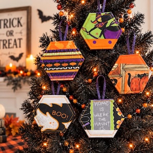 May include: Five hexagonal fabric ornaments hanging on a black Christmas tree. Designs include a witch, orange pumpkins, a white ghost with the text BOO!, and a black sign with the text HOME IS WHERE THE HAUNT IS. Each has a purple ribbon loop.