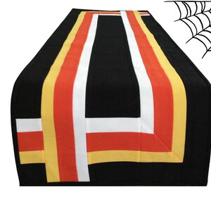 Può includere: Un runner da tavolo nero con strisce gialle, arancioni e bianche. Le strisce sono disposte in un motivo geometrico.