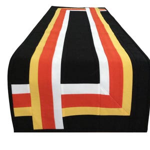 May include: A rectangular table runner featuring a solid black base with a geometric pattern of vertical and horizontal stripes in white, orange, and yellow. The fabric design consists of clean, straight lines that form a rectangular border.