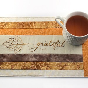 May include: A beige quilted placemat with brown, orange and gold fabric strips. The placemat has an embroidered leaf and the word "grateful" in cursive. A white mug with a brown beverage is on the placemat.
