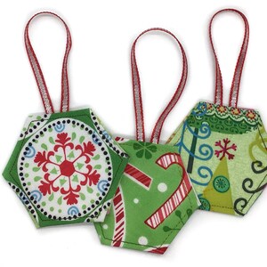 May include: Three hexagonal fabric ornaments with red ribbon loops for hanging. The first features a red snowflake on a white background with green borders. The second is green with red candy canes. The third has a green pattern with swirls.