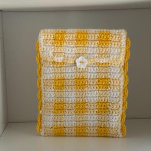 May include: A handmade crochet case in a yellow and white checkered pattern. The case has a scalloped edge and a flap secured with a white flower-shaped button. The case is designed to hold a tablet or e-reader.