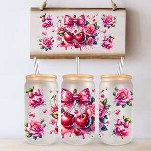 Op de afbeelding: Drie glazen bekers met bamboe deksels en metalen rietjes, versierd met aquarelontwerpen van kersen, roze strikken en rozen. Een houten bord erboven toont vergelijkbare kunstwerken. Het ontwerp heeft een roze en rood kleurenschema.