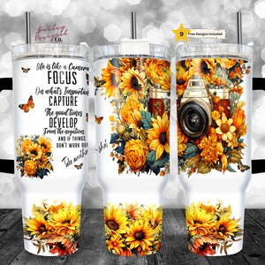 May include: White tumbler with a black handle and a straw. The tumbler features a floral design with sunflowers, butterflies, and a vintage camera. The text on the tumbler reads "Life is like a camera FOCUS on what's important CAPTURE the good times DEVELOP from the negatives AND IF THINGS DON'T WORK OUT Take another shot."