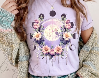 May include: A purple t-shirt with a floral design featuring a full moon surrounded by a circle of crescent moons. The design also includes a dragonfly and a floral wreath.