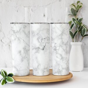 Puede incluir: Tres vasos de acero inoxidable con estampado de m&aacute;rmol blanco y pajitas de pl&aacute;stico transparente. Los vasos est&aacute;n sobre un posavasos de madera.