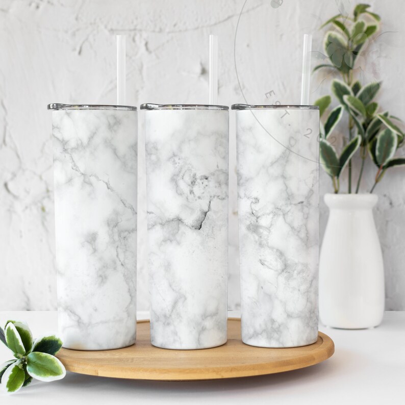 Puede incluir: Tres vasos de acero inoxidable con estampado de m&aacute;rmol blanco y pajitas de pl&aacute;stico transparente. Los vasos est&aacute;n sobre un posavasos de madera.