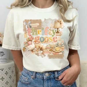 以下が含まれることがあります： 「Thrifty Goose」の文字が入ったコラージュデザインのクリーム色のTシャツ。コラージュにはガチョウ、花、ティーカップ、小さなランプが含まれています。シャツは柔らかい素材で作られています。