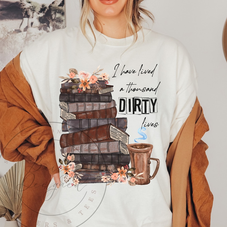 May include: A white t-shirt with a graphic design of a stack of books with flowers and a mug of coffee. The text on the shirt reads "I have lived a thousand DIRTY lives".