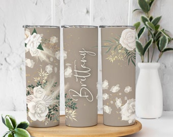 May include: Three personalized stainless steel tumblers with a floral design. The tumblers are beige with white flowers and gold accents. The middle tumbler has the name "Brittany" written on it.