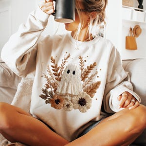 Pode incluir: Camisola creme com um design de fantasma bordado. O fantasma é rodeado por detalhes florais e de folhas em tons de castanho e bege. A camisola tem mangas compridas e gola redonda.