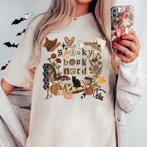 May include: Beige t-shirt with a colorful graphic design featuring the words "spooky book nerd" in a square frame. The design includes a black cat, pumpkins, bats, flowers, and a coffee cup. The shirt is a casual, comfortable style.