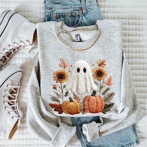 May include: A light gray sweatshirt with an embroidered Halloween design featuring a white ghost, sunflowers, and pumpkins. The sweatshirt is paired with blue jeans and white sneakers, creating a casual fall aesthetic.