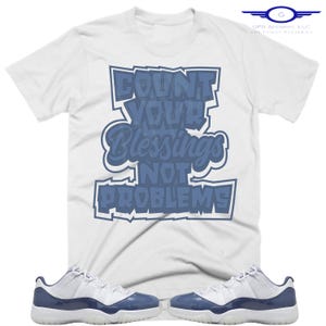 May include: White t-shirt with the text "COUNT YOUR BLESSINGS NOT PROBLEMS" in blue. The shirt is paired with white and navy blue sneakers.