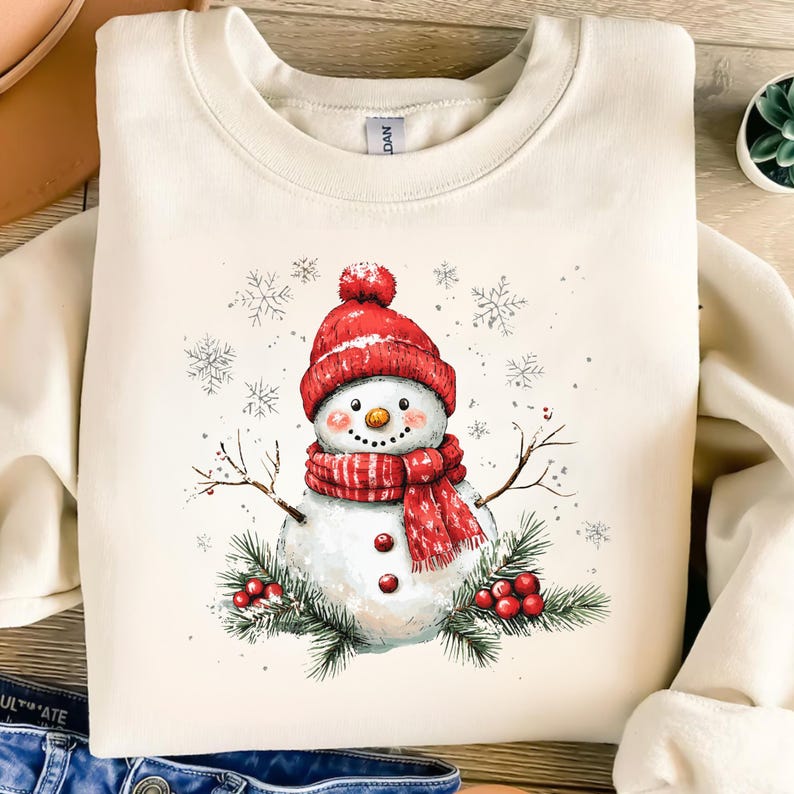Puede incluir: Una sudadera de color crema con un mu&ntilde;eco de nieve de dibujos animados que lleva un gorro y una bufanda rojos. El mu&ntilde;eco de nieve est&aacute; rodeado de copos de nieve y ramas de pino con bayas rojas.