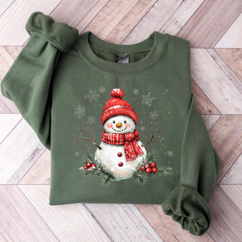 Puede incluir: Una sudadera verde con un dise&ntilde;o de mu&ntilde;eco de nieve. El mu&ntilde;eco de nieve lleva un gorro y una bufanda rojos y tiene una cara blanca con ojos negros y una boca roja. El mu&ntilde;eco de nieve est&aacute; rodeado de bayas rojas y ramas de pino verdes.
