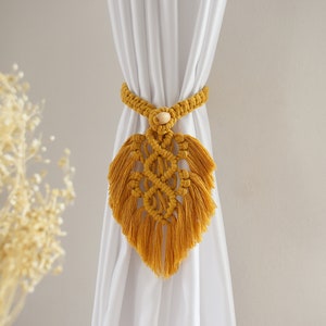 May include: A mustard yellow macrame curtain tieback with a wooden bead in the center. The tieback is shaped like a heart and has fringe details.