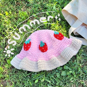 K&ouml;nnte beinhalten: Ein rosa-wei&szlig;er H&auml;kel-Sonnenhut mit Erdbeer-Design. Der Hut liegt auf einem Bett aus gr&uuml;nem Gras.