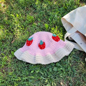 K&ouml;nnte beinhalten: Ein rosa geh&auml;kelter Bucket-Hut mit wei&szlig;em Rand und drei gestickten Erdbeer-Designs.