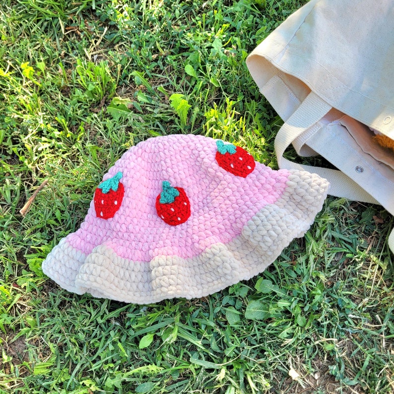 K&ouml;nnte beinhalten: Ein rosa und wei&szlig;er geh&auml;kelter Bucket-Hut mit Erdbeer-Applikationen. Der Hut hat eine breite Krempe und einen gekr&auml;uselten Rand.
