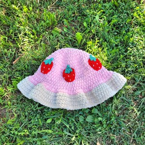K&ouml;nnte beinhalten: Ein rosa geh&auml;kelter Bucket-Hut mit wei&szlig;em Besatz und drei gestickten Erdbeermotiven.