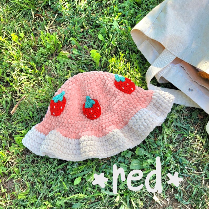 K&ouml;nnte beinhalten: Ein rosa geh&auml;kelter Bucket-Hut mit wei&szlig;em Rand und gestickten Erdbeermotiven. Der Hut liegt auf einem Bett aus gr&uuml;nem Gras.
