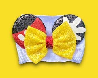 May include: A white headband with black and red Mickey Mouse ears and a yellow bow. The ears are covered in sequins.