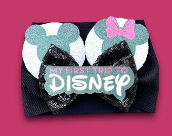 May include: A black headband with two Mickey Mouse ear designs, one with a pink bow. The ears are white and teal glitter. A black sequin bow with the text "MY FIRST TRIP TO DISNEY" is centered on the headband.