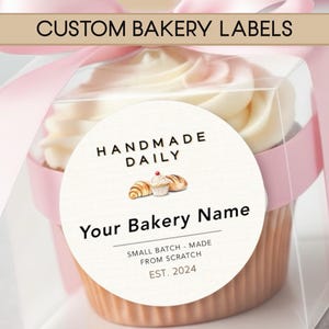 May include: A cupcake in a clear box with a pink ribbon. The cupcake has white frosting and a round label that says "HANDMADE DAILY" and "Your Bakery Name". The label also includes the text "SMALL BATCH - MADE FROM SCRATCH EST. 2024". The top banner says "CUSTOM BAKERY LABELS".