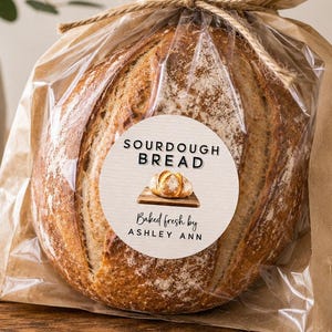 May include: A round loaf of sourdough bread packaged in a clear plastic bag inside a brown paper bag, tied with a jute twine bow. A circular white label on the front reads SOURDOUGH BREAD, with an illustration of bread and Baked fresh by ASHLEY ANN.