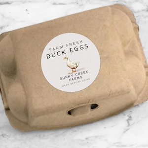 May include: A brown cardboard egg carton with a round label. The label reads "Farm Fresh Duck Eggs" and features a watercolor illustration of a duck. The text "Sunny Creek Farms" and "Wash Before Using" are also on the label.