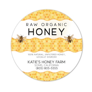 May include: A round label with the words "RAW ORGANIC HONEY" in black. The label features a honeycomb design and two illustrations of bees. The text "100% NATURAL, UNFILTERED HONEY, LOCALLY SOURCED" is also included. The label also includes the text "KATIE'S HONEY FARM SOMIS, CALIFORNIA (805) 805-5555".
