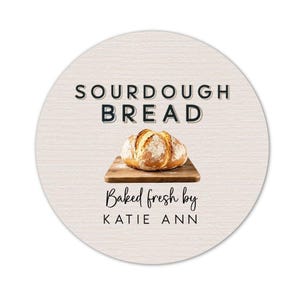 May include: Round sticker with the words "SOURDOUGH BREAD" above an image of a loaf of bread on a wooden cutting board. The text "Baked fresh by KATIE ANN" is below the bread image.