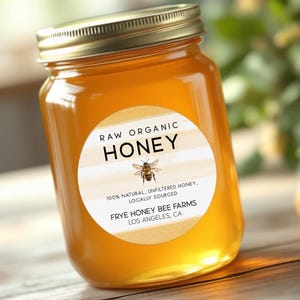 May include: A glass jar filled with golden honey, topped with a gold lid. The jar has a round label with the words "RAW ORGANIC HONEY" and a bee illustration. The label also states "100% NATURAL, UNFILTERED HONEY, LOCALLY SOURCED" and the name of the farm.