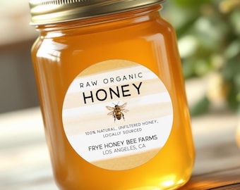 May include: A glass jar filled with golden honey, topped with a gold lid. The jar has a round label with the words "RAW ORGANIC HONEY" and a bee illustration. The label also states "100% NATURAL, UNFILTERED HONEY, LOCALLY SOURCED" and the name of the farm.
