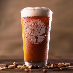May include: A pint glass filled with amber beer, topped with a creamy white head. The glass is branded with "Zombie Brew" and "Gluten Free" text. Condensation beads on the glass. Mixed nuts are scattered on a wooden surface at the base.