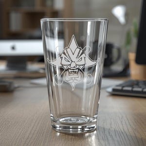 May include: A clear pint glass with a white graphic of a demon face. The glass is empty and stands on a wooden desk. The background includes a computer monitor, keyboard, and other office items.