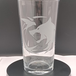 May include: Clear pint glass featuring an etched wolf's head design. The glass has a cylindrical shape with a flared top. The wolf design is surrounded by stylized elements. The glass is made of clear glass.